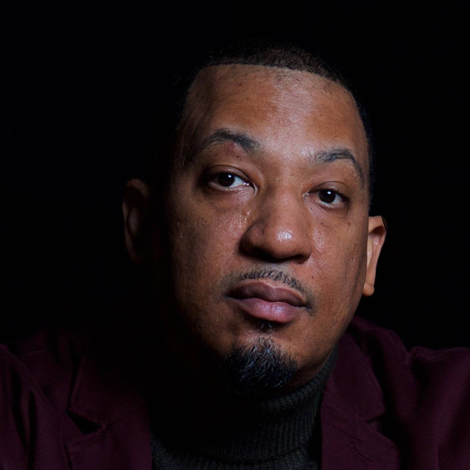 A portrait of Dasan Ahanu in front of a black background. He wears a turtleneck and blazer.