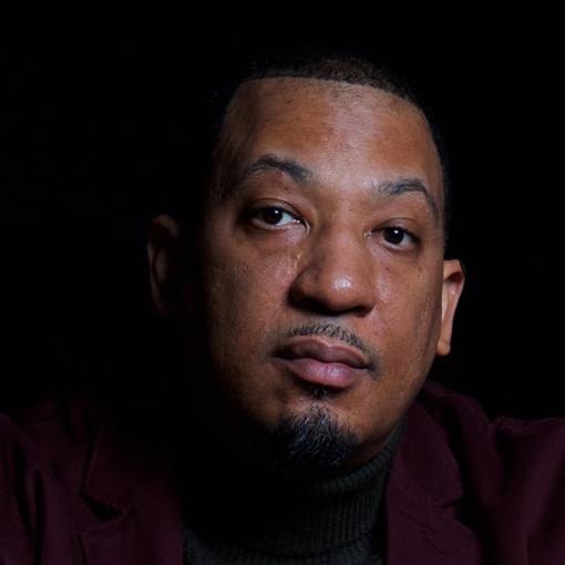 A portrait of Dasan Ahanu in front of a black background. He wears a turtleneck and blazer.