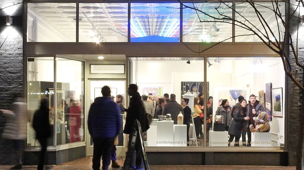 A nighttime view of a crowd inside a gallery from the outside.