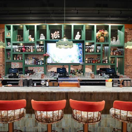 View of the bar at Primrose Bar and Lounge with bar stools and a mirrored back bar and green fringed chandeliers.