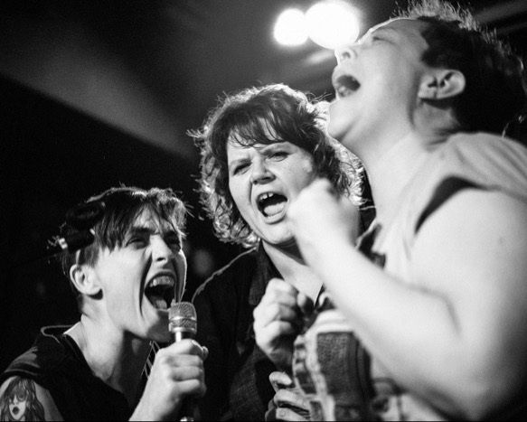 A black and white photo depicts 3 people on stage singing energetically into microphones.