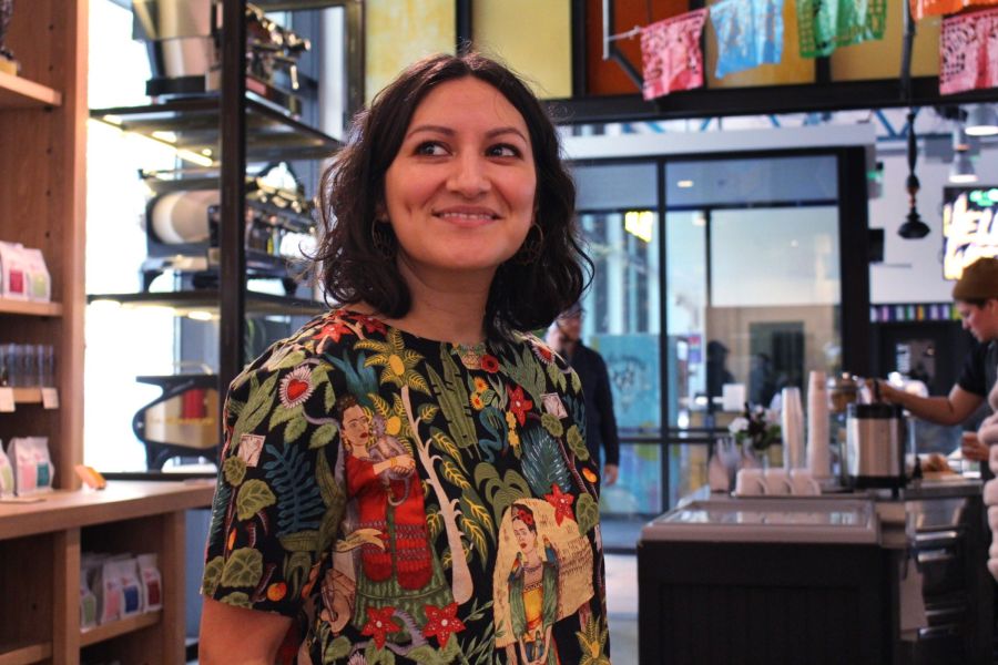 A woman smiles in front of the counter of a coffee shop and coffee roasting equipment.