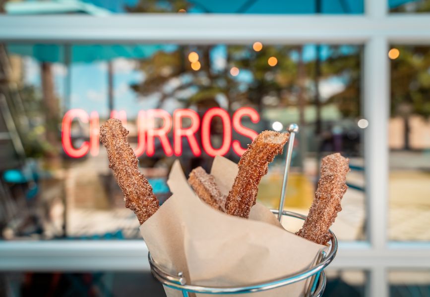 A neon "Churros" sign is eclipsed by a batch of the confection. Cocoa Cinnamon Lakewood & 4th Dimension Coffee Roastery.