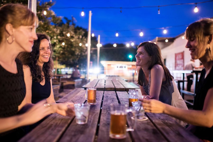 A group of women enjoys drinks around an outdoor table in the evening with bistro lights overhead.