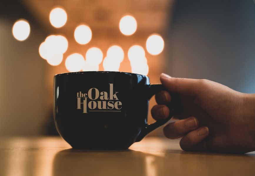 With lights out of focus in the background creating a dreamy view, a customer holds a mug with The Oak House's logo on it.