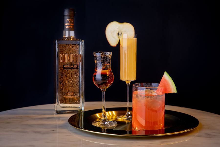 A bottle of conniption Gin stands next to two colorful cocktails on a table.