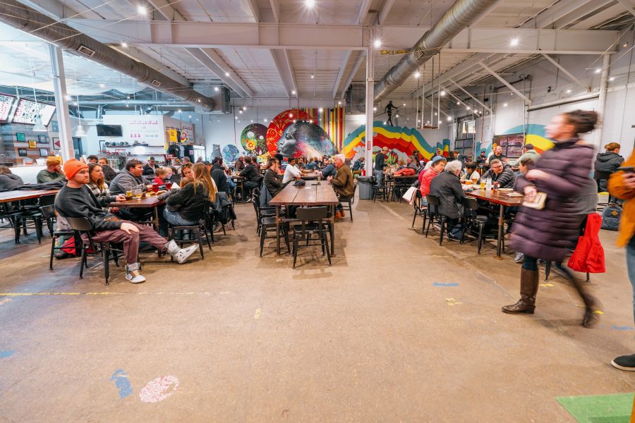 A crowd of people sit at long tables and drink beer in front of a colorful mural on the back wall of the brewery.