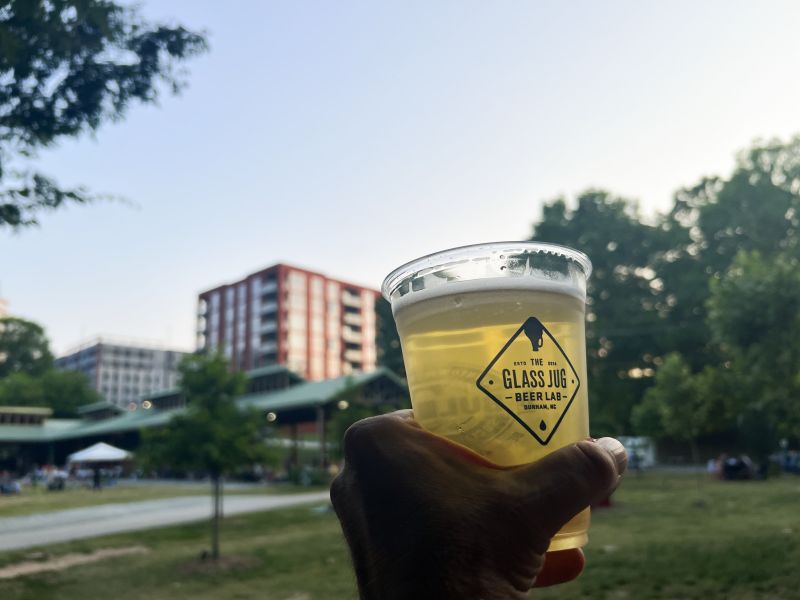 A hand holds a beer from The Glass Jug in the air in front of the Durham Central Park overhang.
