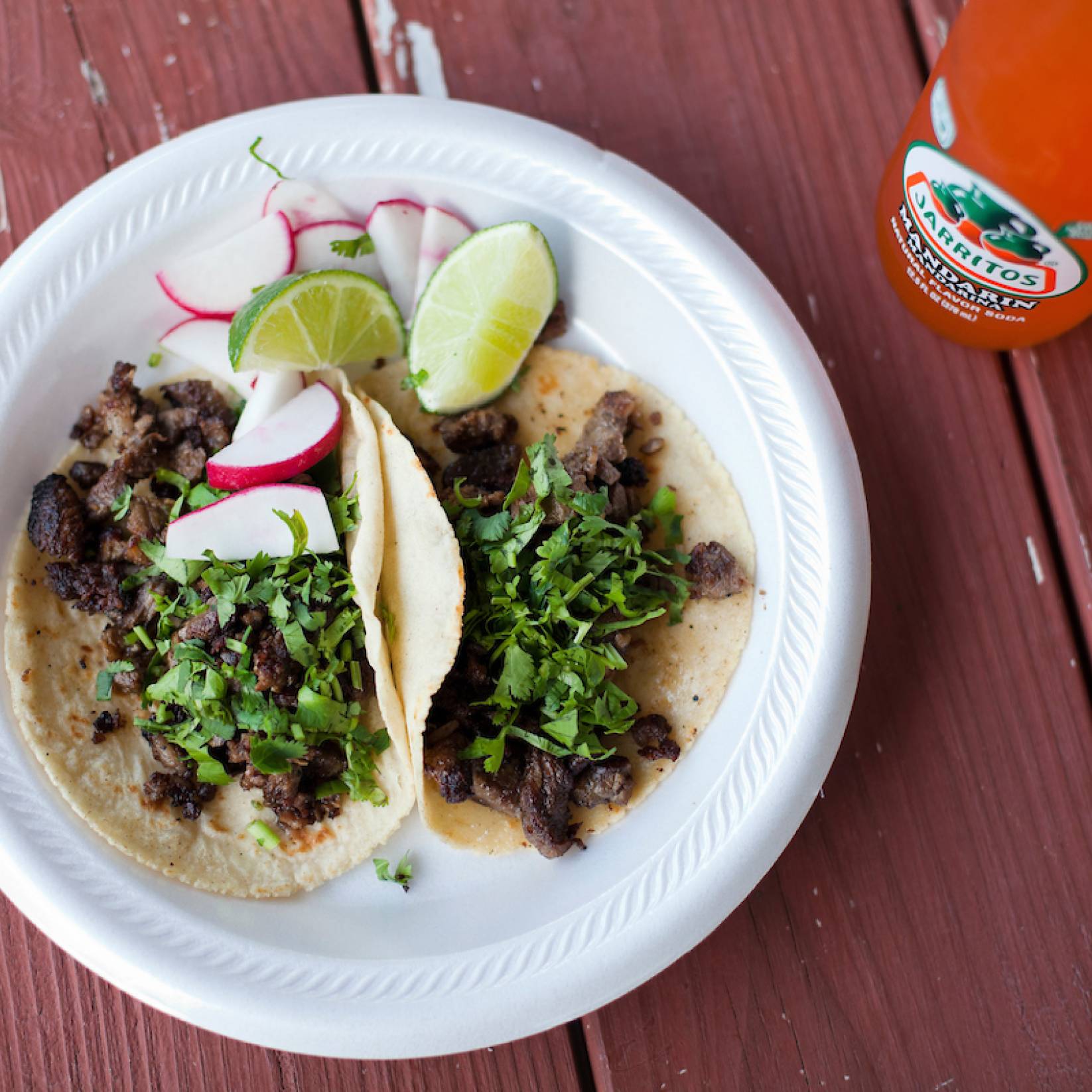 A pair of tacos sit on a plate next to limes and radishes on a picnic table.