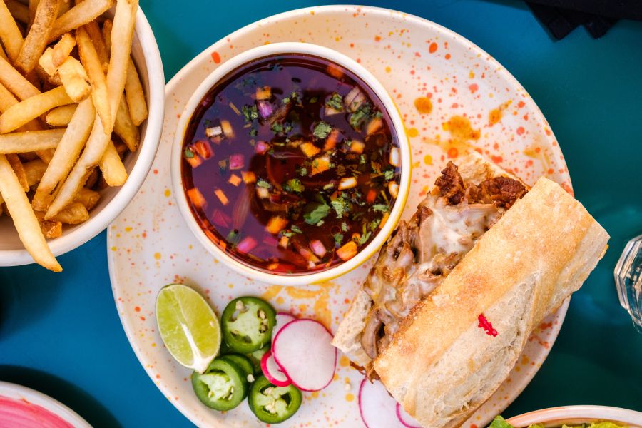 A birria dip sandwich sits on a plate next to a fresh bowl of fries at Queeny's in Durham, NC.