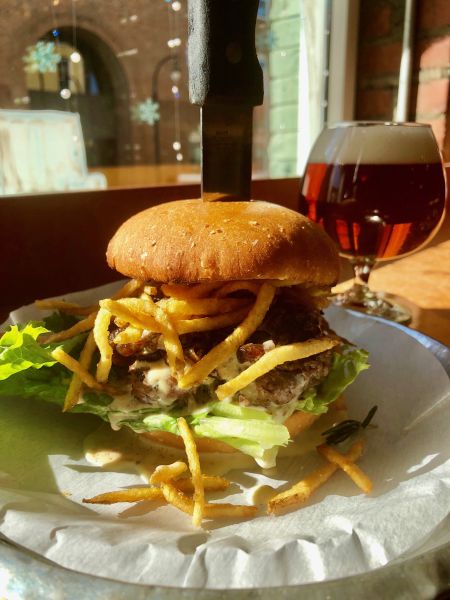A burger is stacked high on a plate held together by a knife with an amber beer sitting in the background.