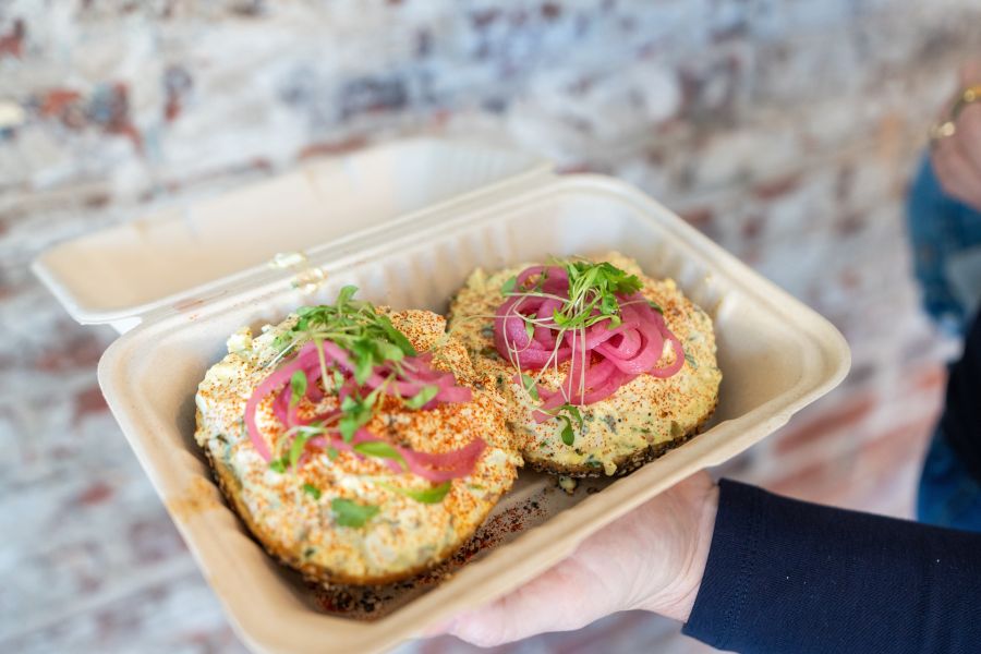 A large bagel from Isaac's Bagels is depicted with a generous amount of spread, spices, and pickled onions.