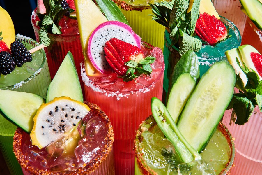 Green, yellow and pink cocktails from TaTaco sit on a table garnished with colorful fruit and vegetables.