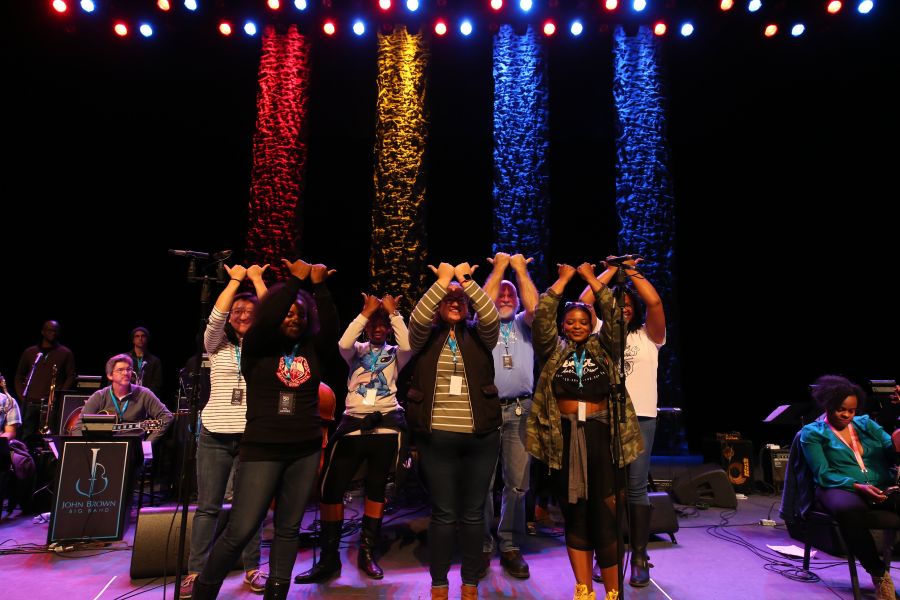 A group of employees stand and bow at a closing ceremony at Durham Performing Arts Center under Durham flag colored lights.