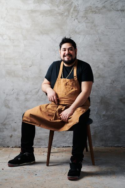 Chef Oscar Diaz wears a black shirt and brown apron while he sits on a chair and smiles.