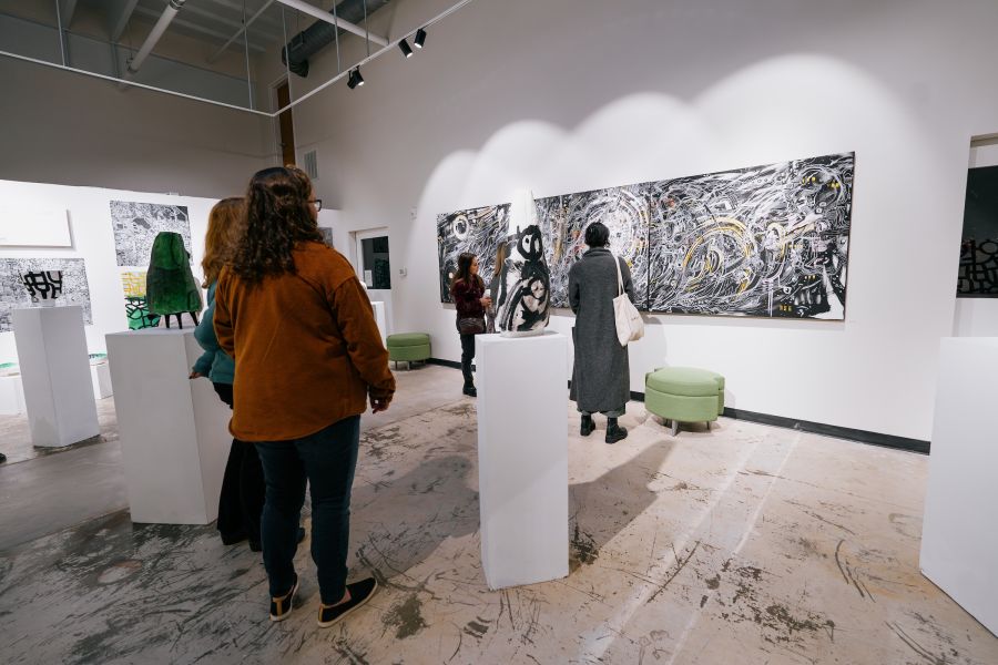 People view art on walls and pillars in the Durham Art Guild Gallery. The floor is marked with paint and art supplies.