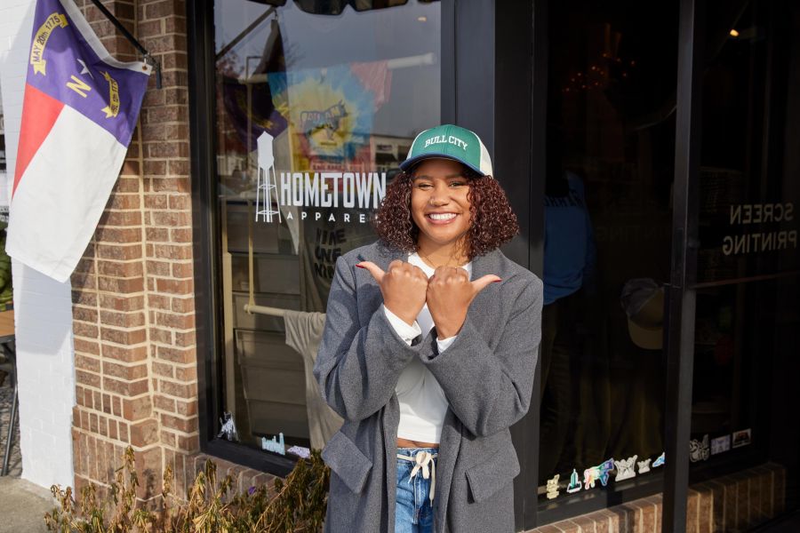 A woman stands outside of the storefront Hometown Apparel on Durham's Ninth Street and makes a bull symbol with her hands