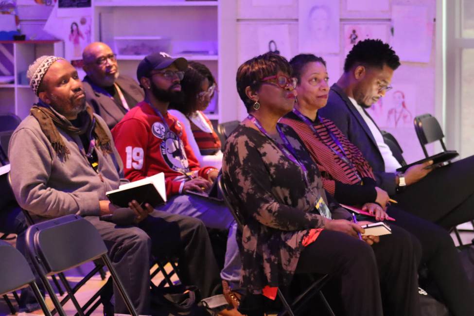A crowd of attendees sits in rows of chairs and listens to a speaker, out of frame.