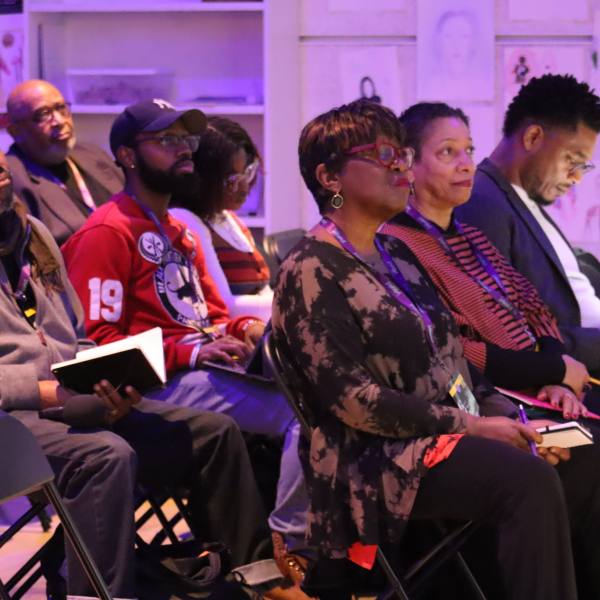 A crowd of attendees sits in rows of chairs and listens to a speaker, out of frame.