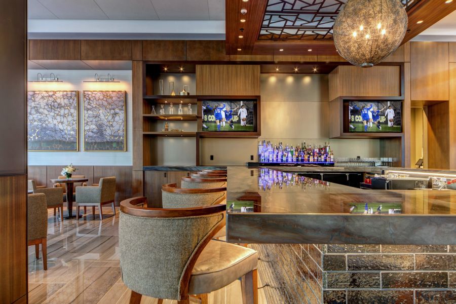 A bar with grey upholstered stools and televisions playing sports in the background. A lounge area with chairs to the left.