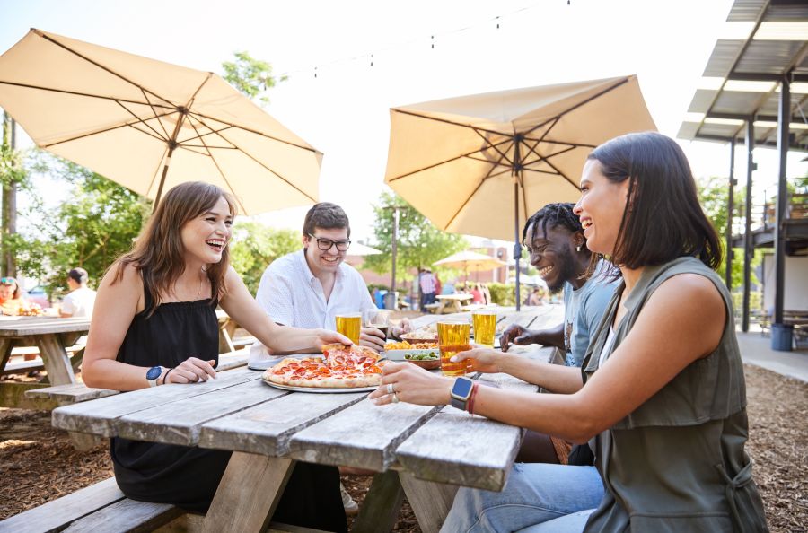 4 friends laugh together while enjoying pepperoni pizza and beers in the sun at Ponysaurus Brewing Co. in Durham.