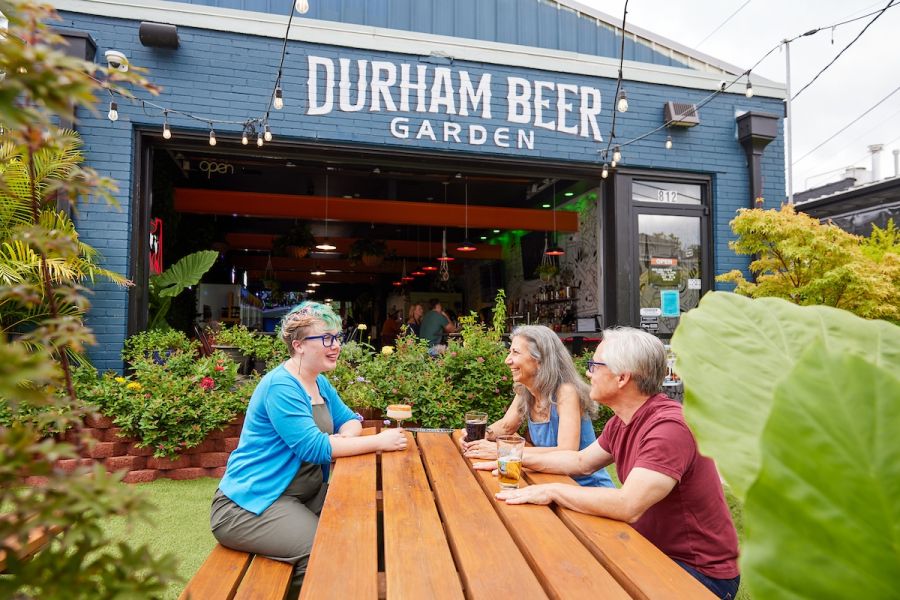 Three people enjoy beers while sitting at a picnic table on a lawn in front of a building with a Durham Beer Garden sign.