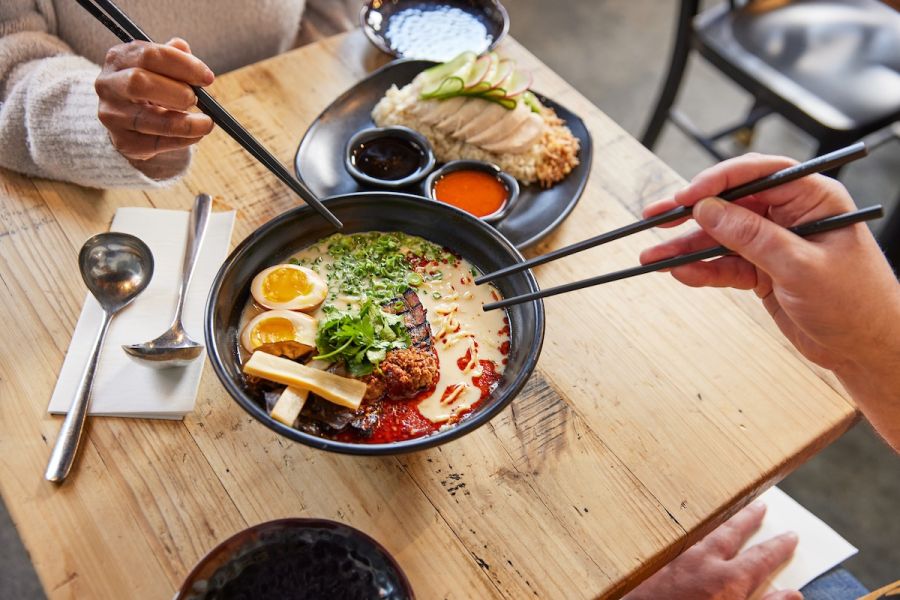 Two diners reach toward a bowl of ramen with chopsticks from across a table. A dish with chicken and sauces is on the table.