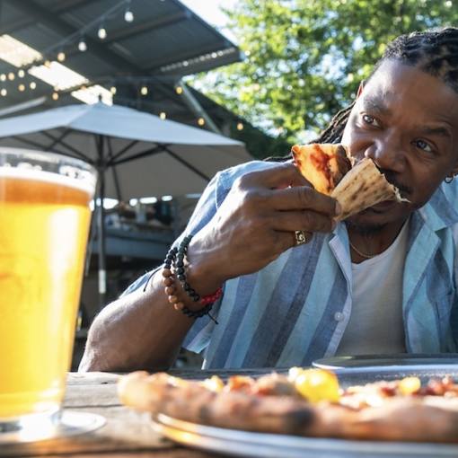A man sits at an outdoors picnic table at Ponysaurus biting into a slice of pizza with a beer next to him.