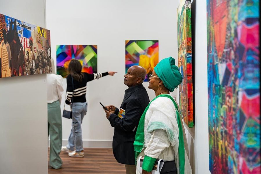 A man and a woman look at colorful art on a gallery wall. In the background, a woman points at a painting on the wall.