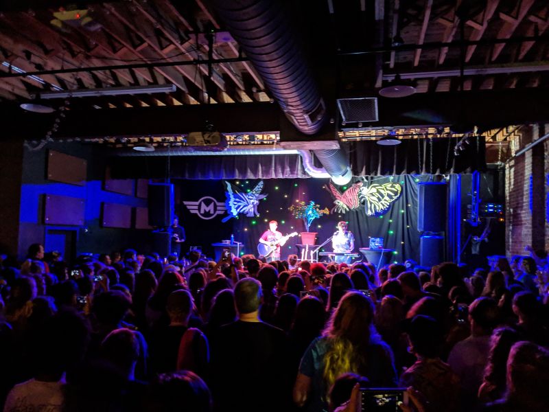 Concert goers stand behind a lit stage at a live performance at Motorco Music Hall.