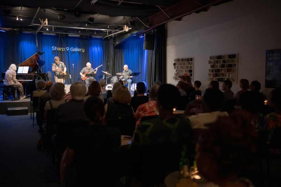 A band plays on stage for a crowd at Sharp 9 Gallery as the audience looks on.