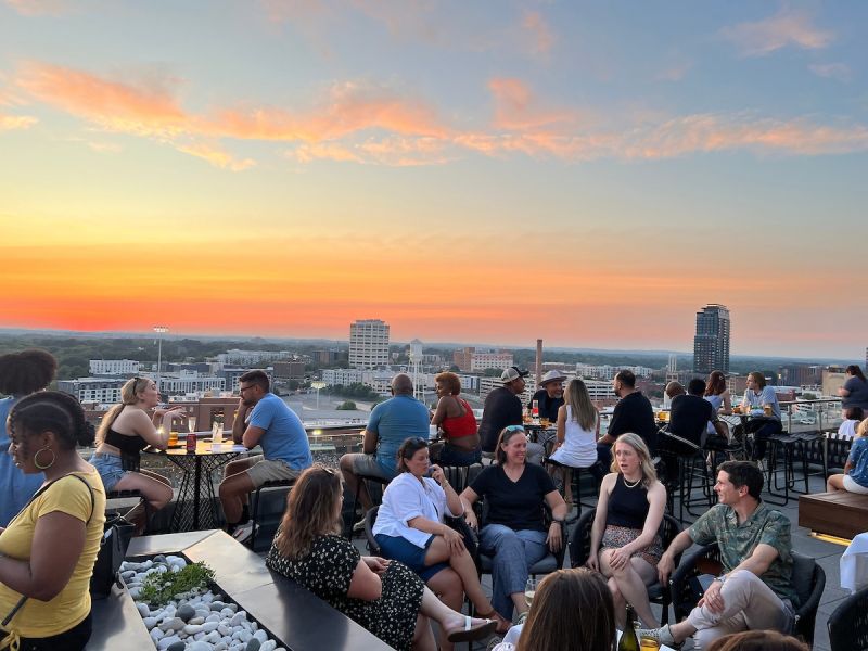 A crowd of people sit at tables at The Lenny's rooftop patio overlooking the Durham Bulls Athletic Park and downtown Durham.