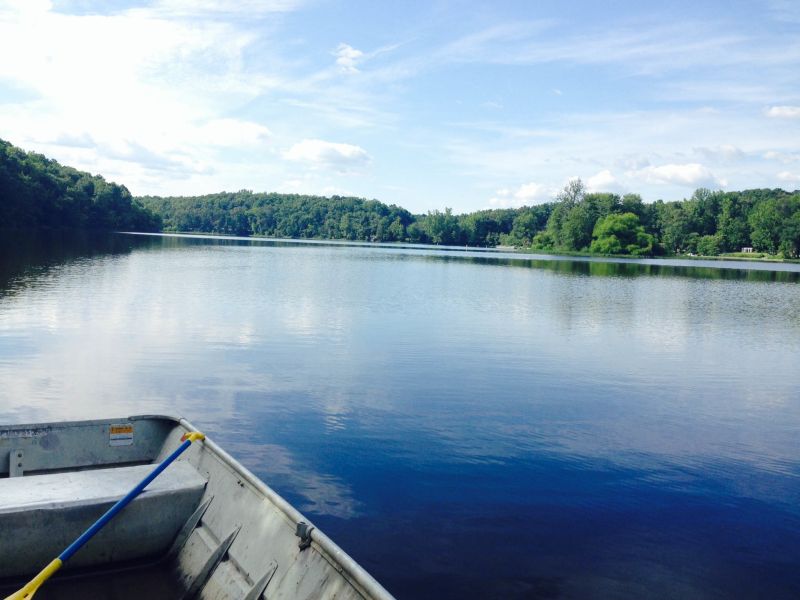 A paddle rests in the corner of a flat-bottom aluminum boat floating over a serene and sunny Lake Michie.
