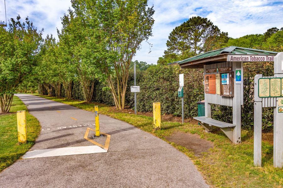 A paved trail lined by trees. Trailhead signage says American Tobacco Trail.
