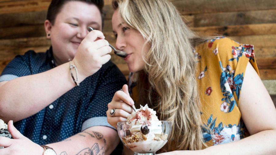 A couple shares an ice cream sundae. One person feeds the other with a spoon.