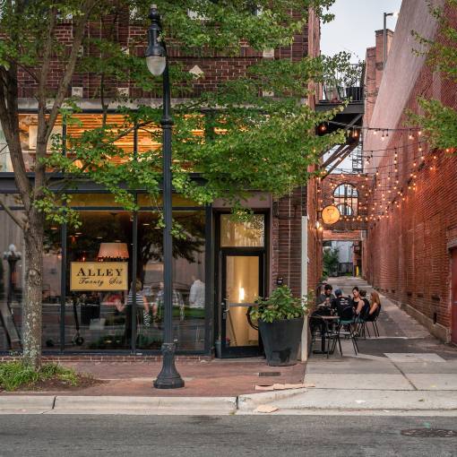 A photo of a restaurant in downtown from across the street. People dining inside and outside in the alley next door.
