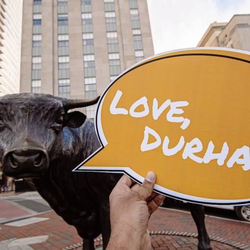 A speech bubble that says &quot;Love, Durham&quot; being held in front of the Major the Bull statue. City streets in the background.