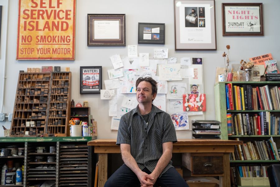 A man at Horse & Buggy Press sits in front of a desk facing the camera and smiling. Art and prints line the wall behind him.