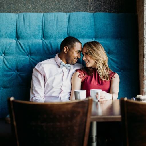 A couple cuddles in a booth with cups of coffee in front of them.
