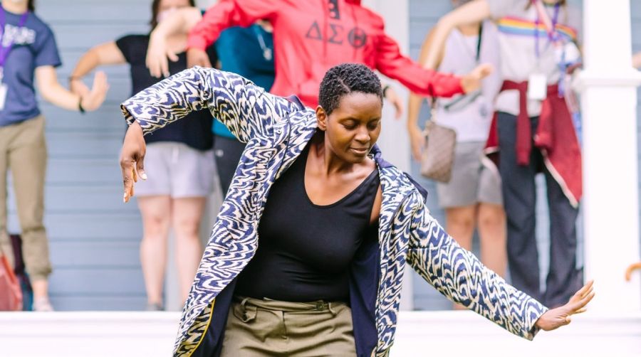 A woman leads a group of people in a dance. Her arms are outstretched as she leans forward and looks down.